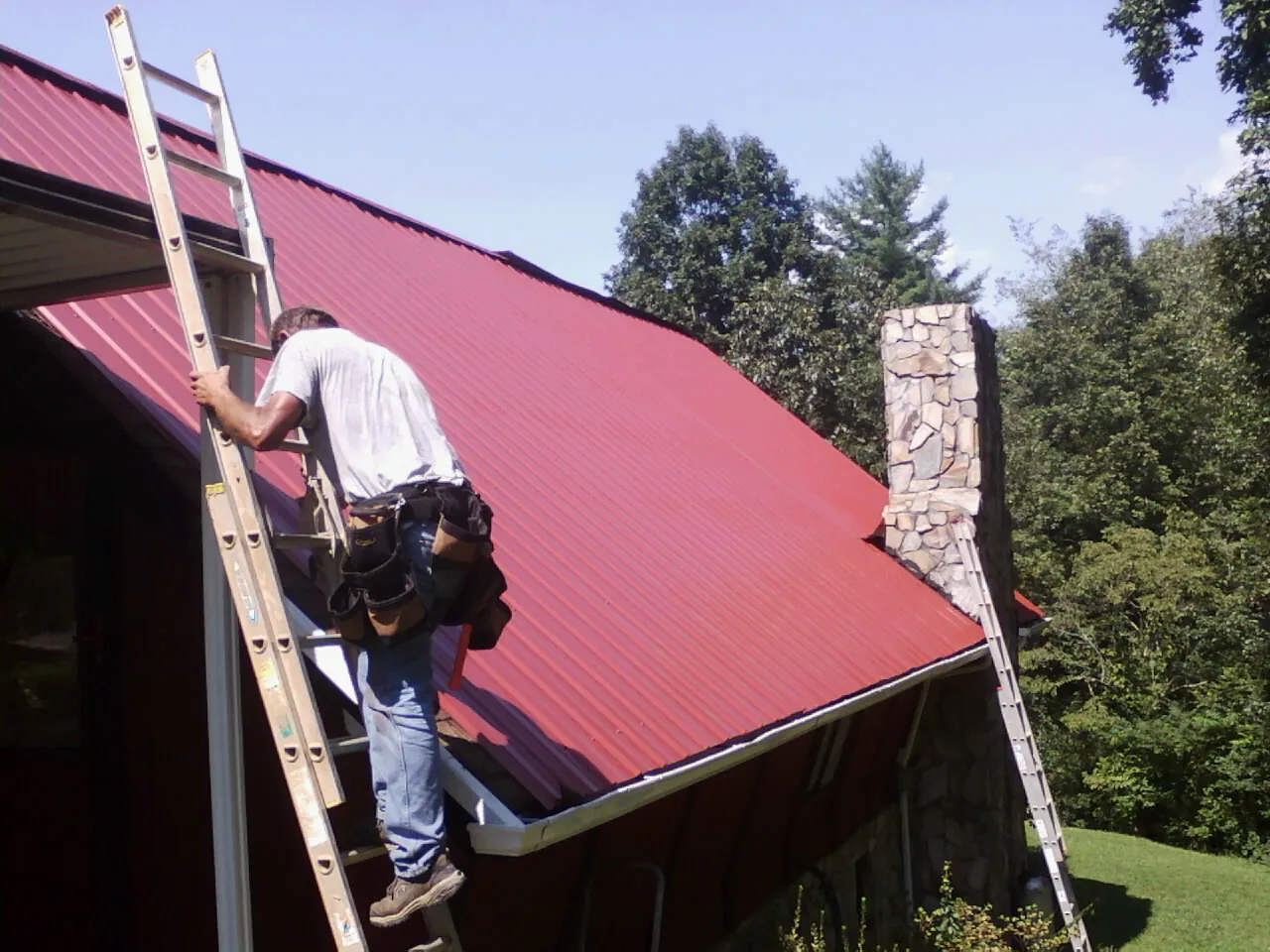 Roofer on a ladder beside a red metal roof and stone chimney — Soffit & Fascia Repair by Roofing Brothers in Howell